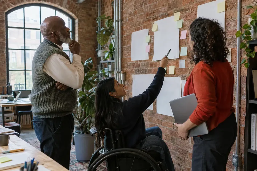 Three colleagues in a brick-walled office at a wall of blank sheets and sticky notes; one, in a wheelchair, points up with a marker.