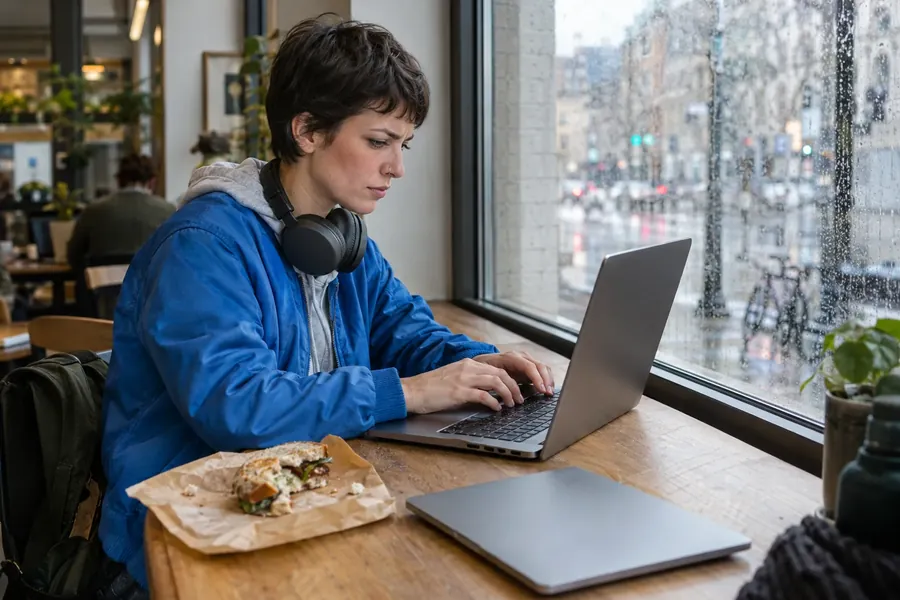 A woman with a short dark haircut and a blue jacket typing on a laptop at a rain-streaked café window, headphones round her neck.